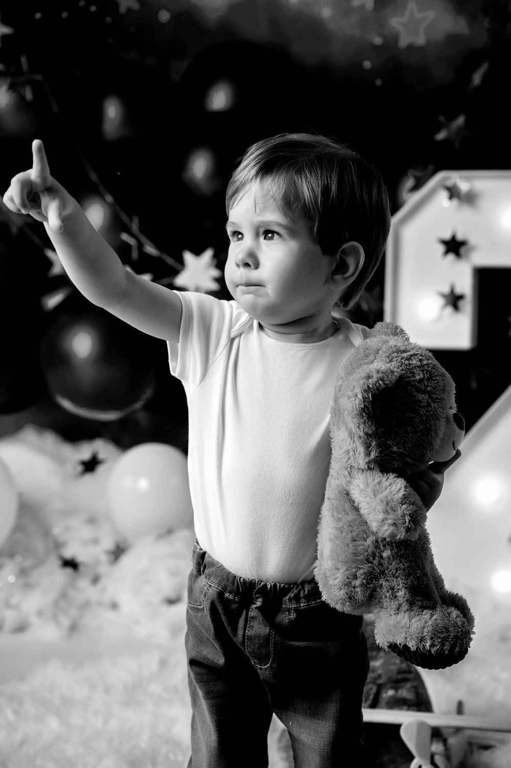 A young child in a white shirt points upward while holding a teddy bear against a starry backdrop.