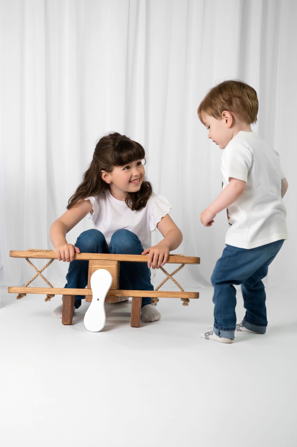 Two children having fun as one sits on a wooden folding stool while the other stands nearby in white shirts.