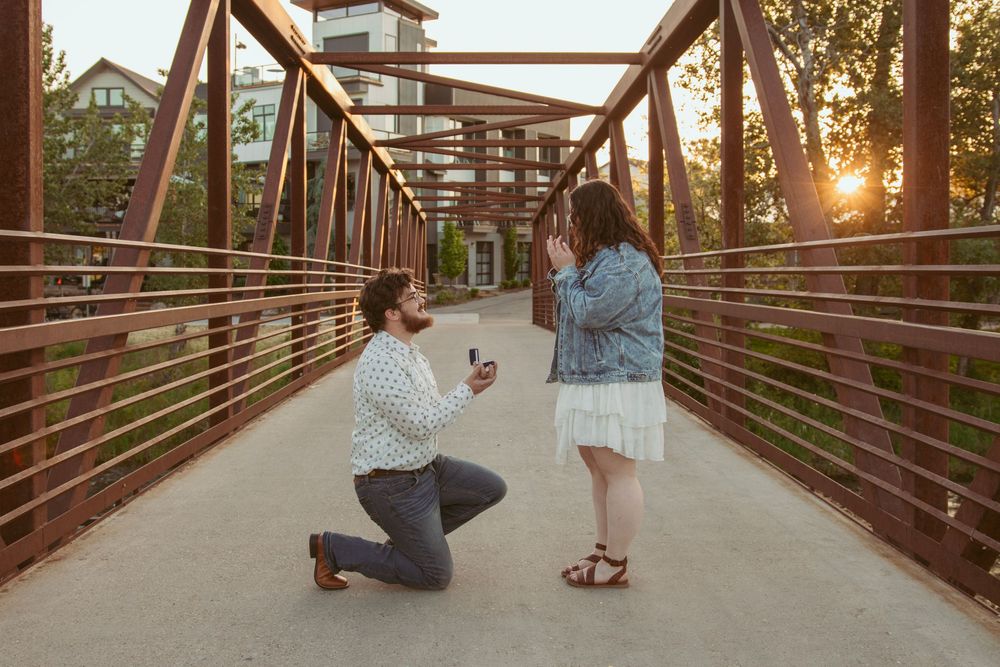 Proposal moment captured at sunset on rustic metal bridge as someone kneels with engagement ring.