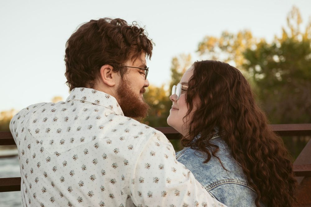 A couple in casual attire share an intimate moment at sunset on a deck while wearing a patterned sweater and denim jacket.