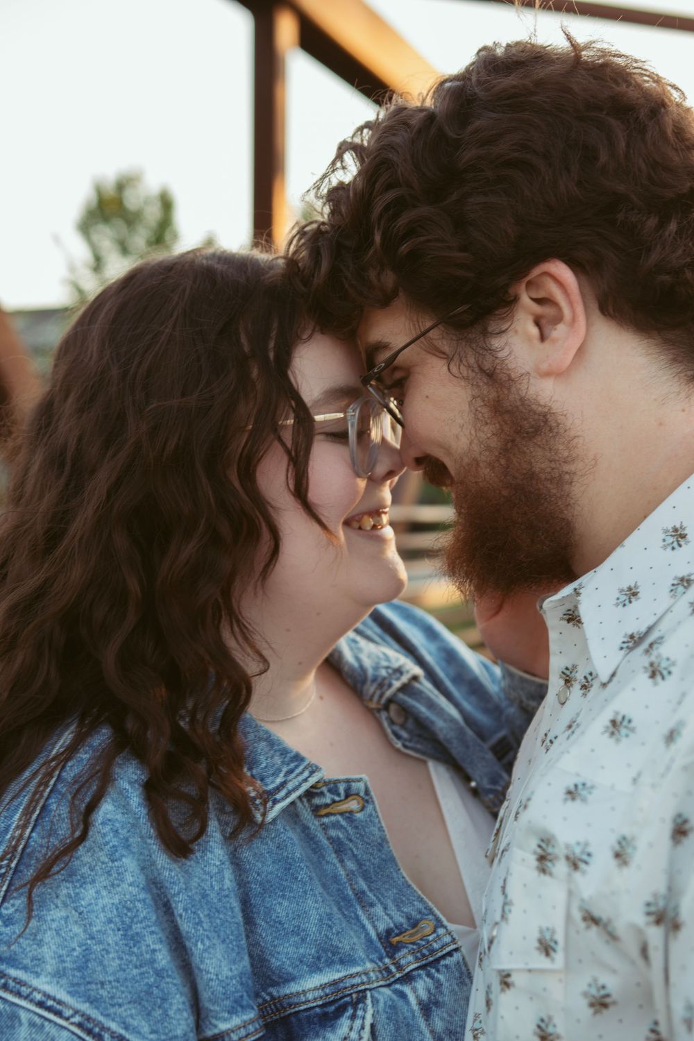 Romantic couple in glasses share an intimate moment outdoors, with one wearing denim and the other a floral print.