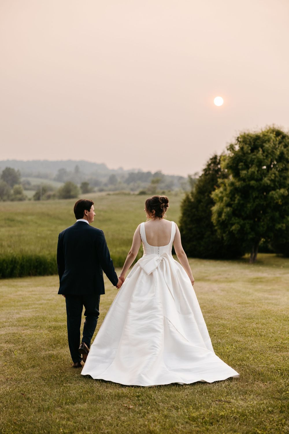 Newlywed couple walks into sunset across rural meadow with bride's white wedding dress trailing behind.