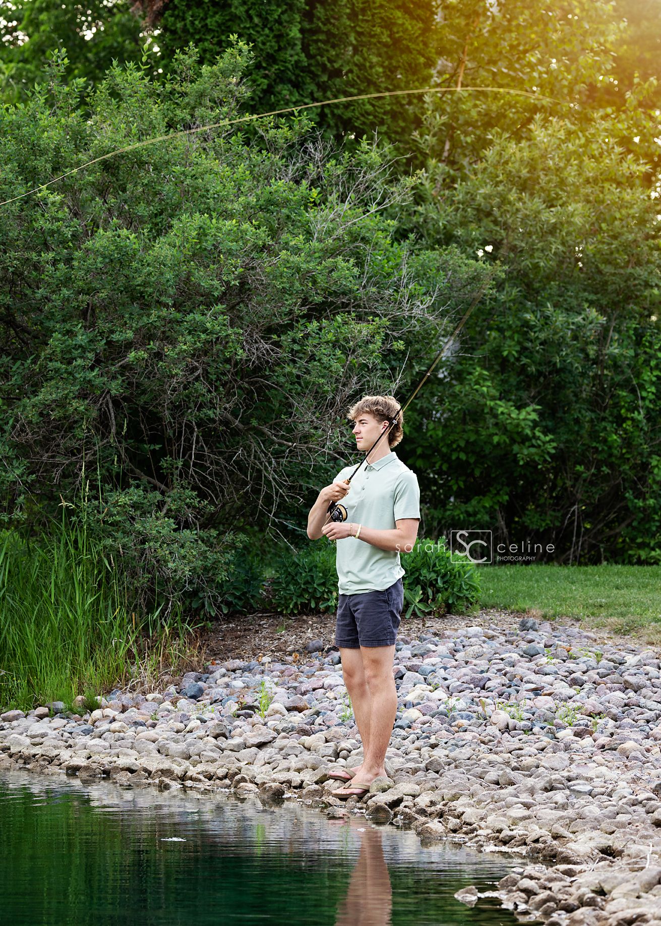 A person holding a fishing rod stands on rocky shore of a pond during golden hour with lush greenery in background.