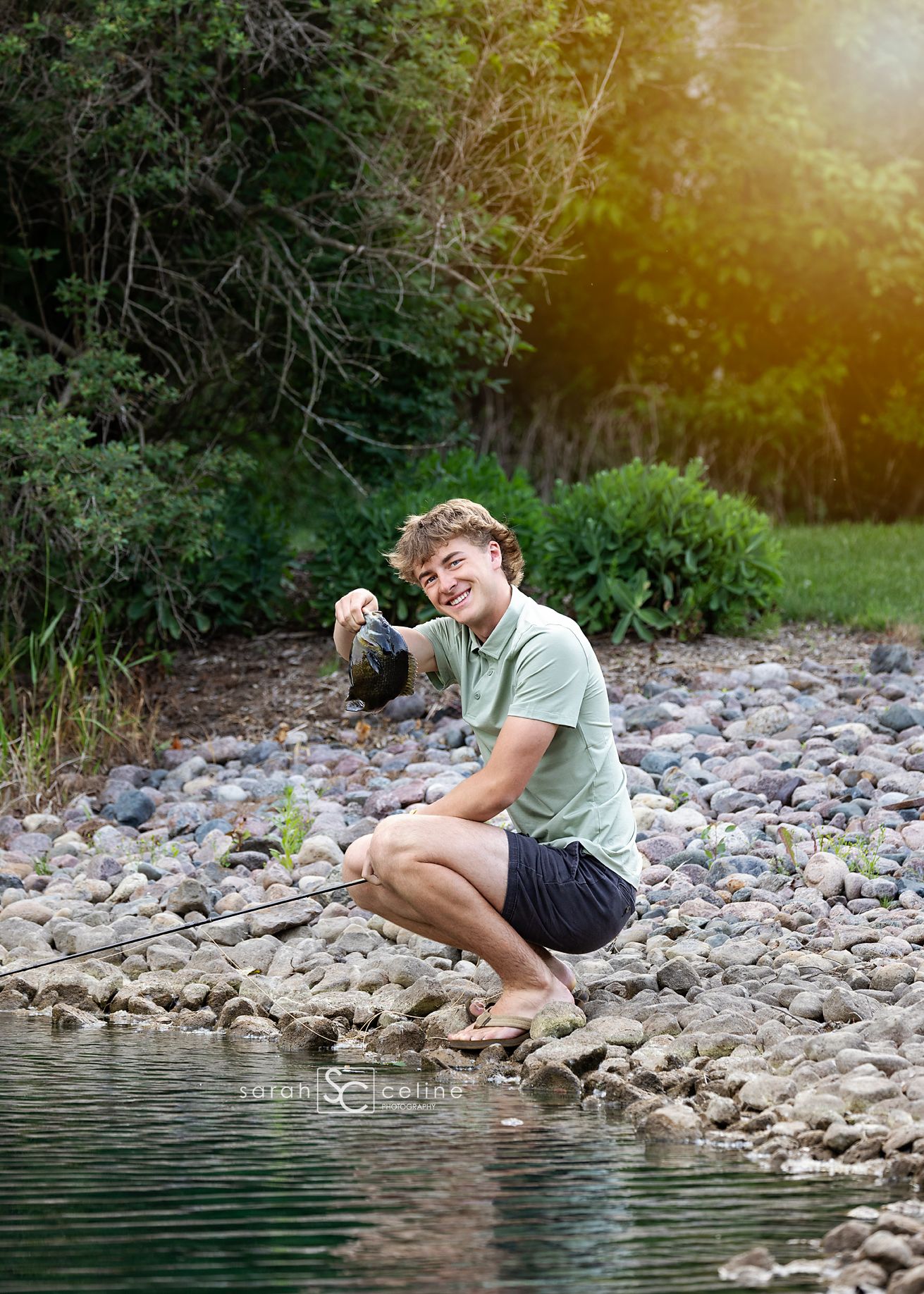 Someone in casual attire crouches on rocky riverbank while taking photos near water and trees in warm sunlight.