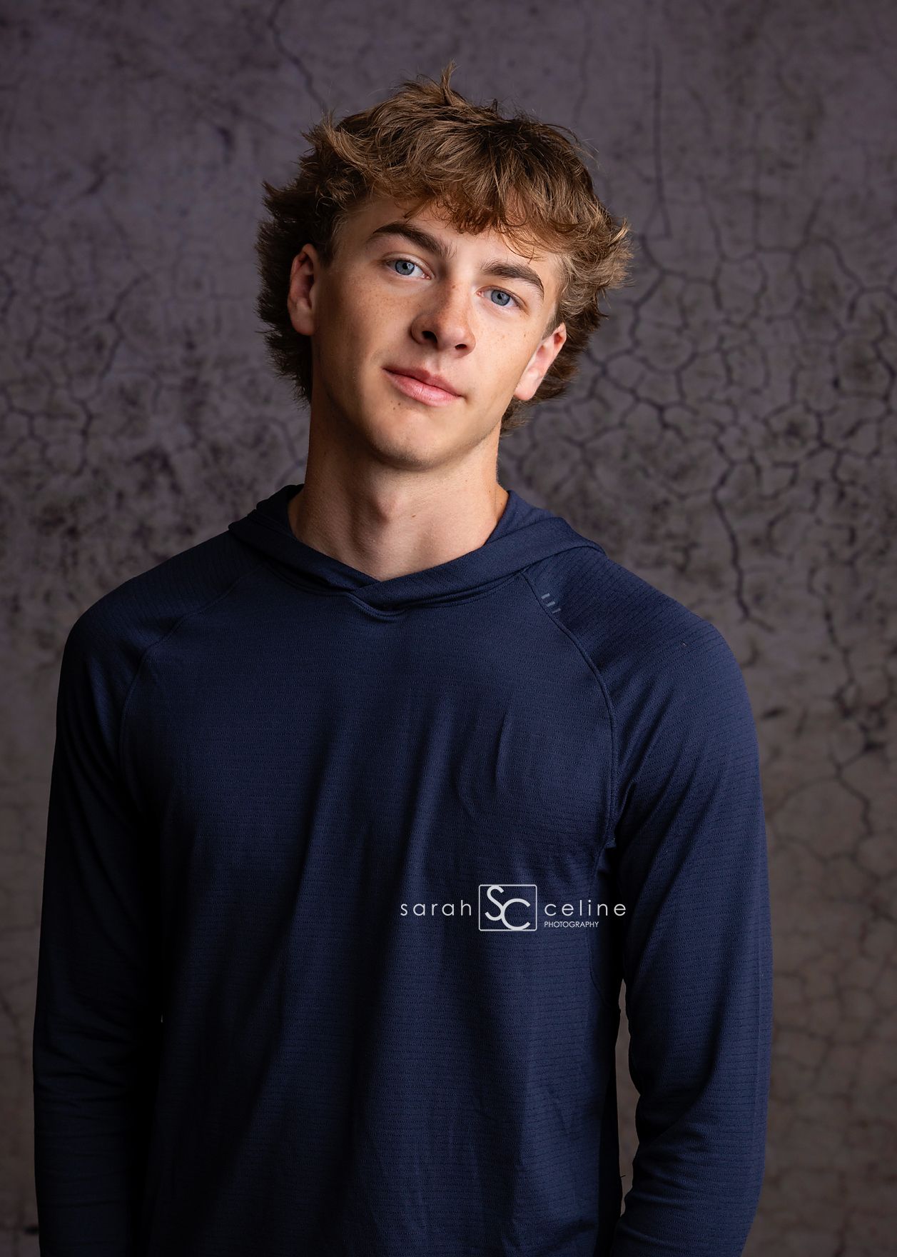 Portrait shot of a person wearing a navy crew neck shirt with a company logo against a dark textured background.