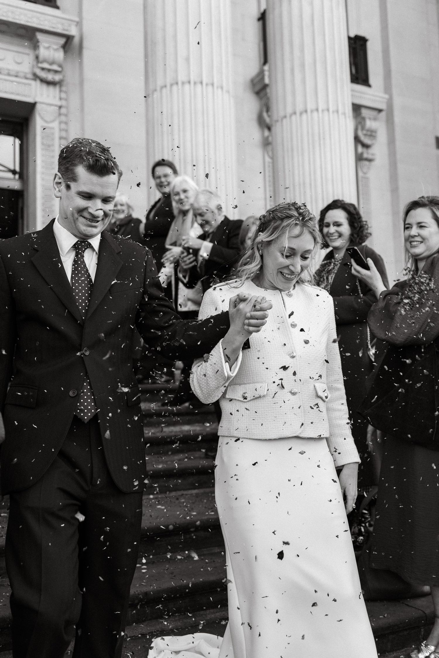 Wedding celebration outside a columned building with confetti falling in black and white.