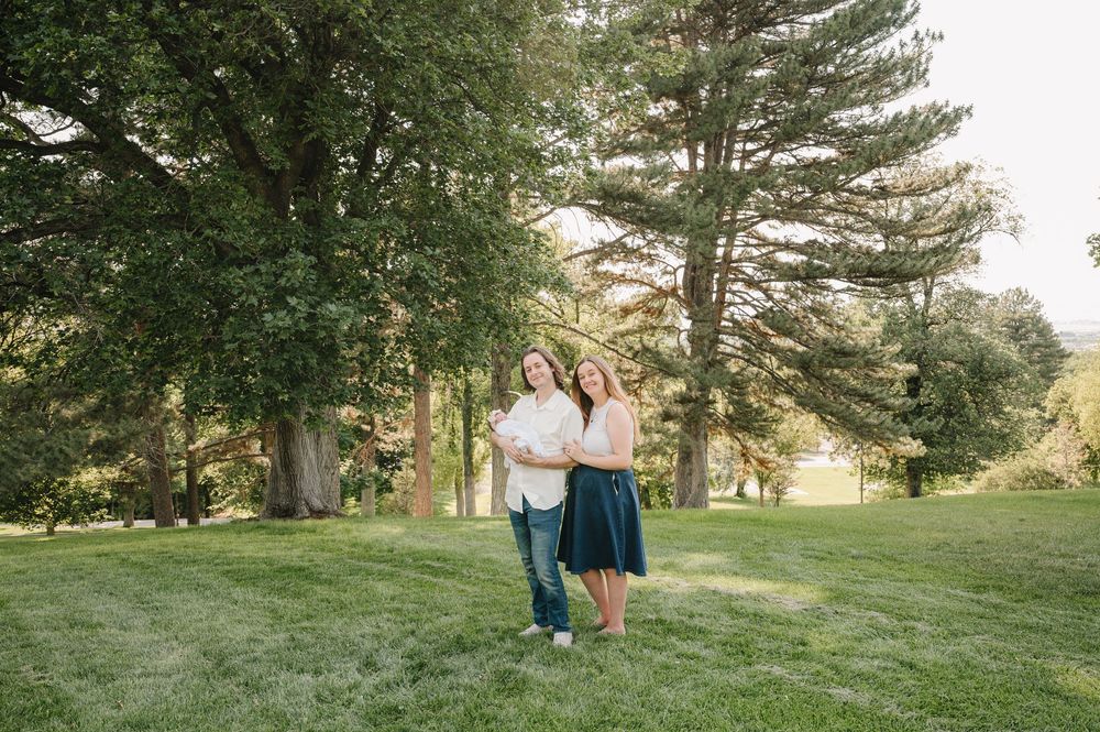 Couple smiling together on lush green lawn surrounded by tall trees in a sunny park setting.