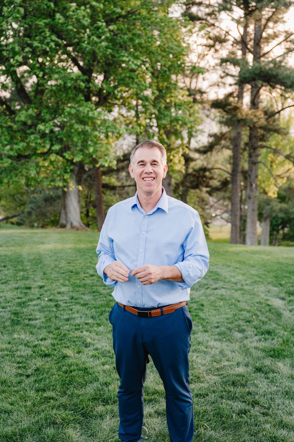 Smiling man in blue shirt and navy pants standing on green grass with tall trees in background.