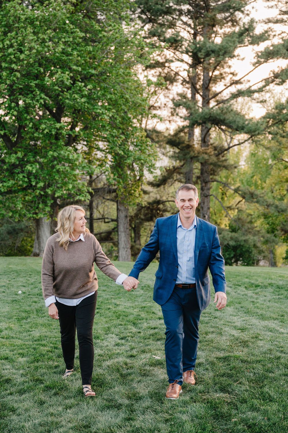 A smiling couple walks hand-in-hand through a lush green park, man in blue blazer, woman in tan sweater.