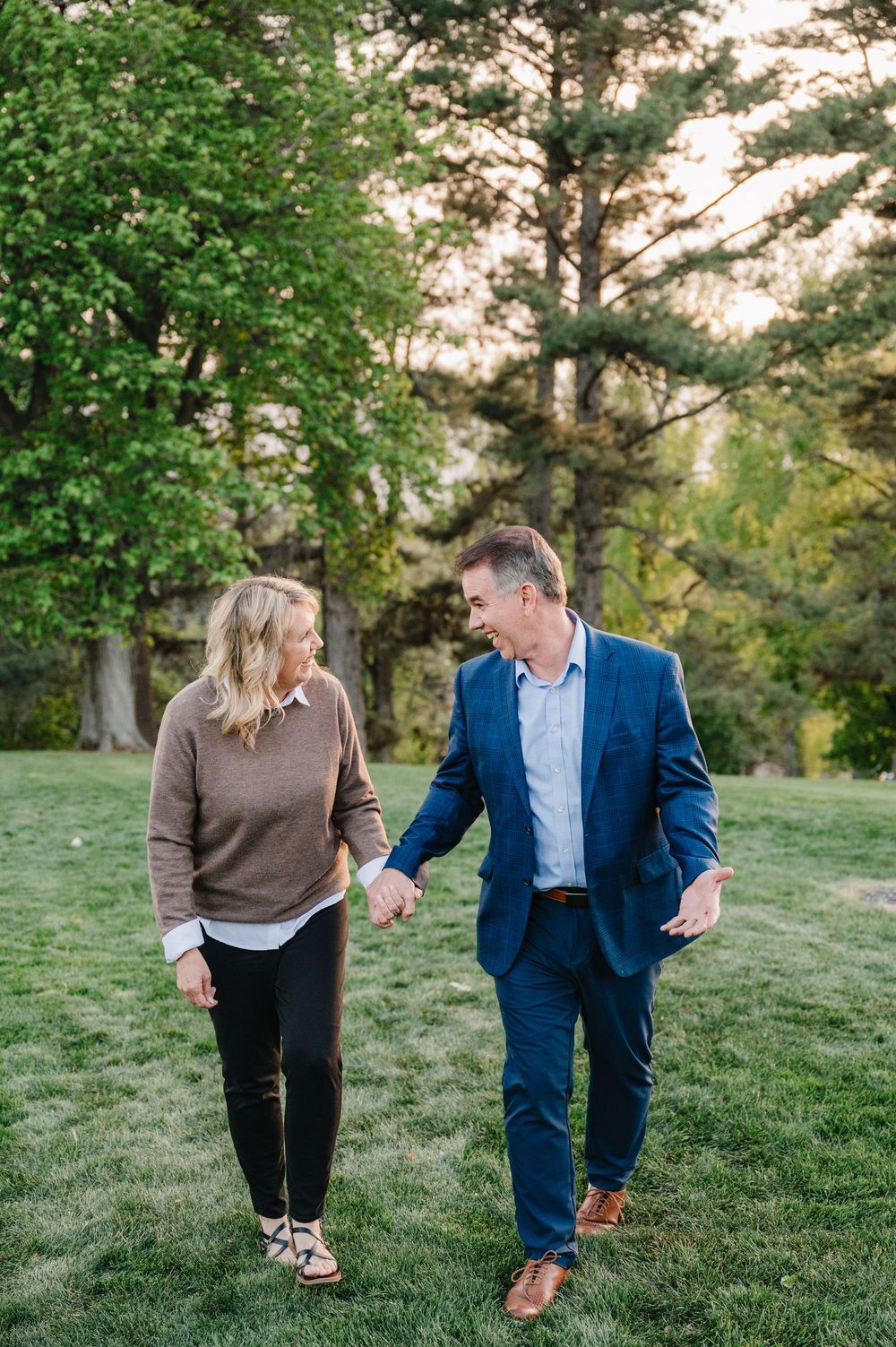 A couple holds hands and smiles while walking through a lush green park, woman in brown sweater, man in blue suit.