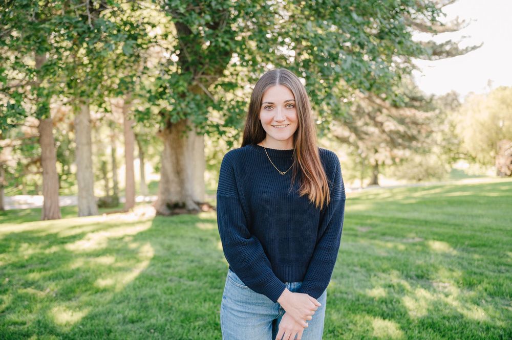 Young woman in navy sweater smiling outdoors on green lawn with trees in background.