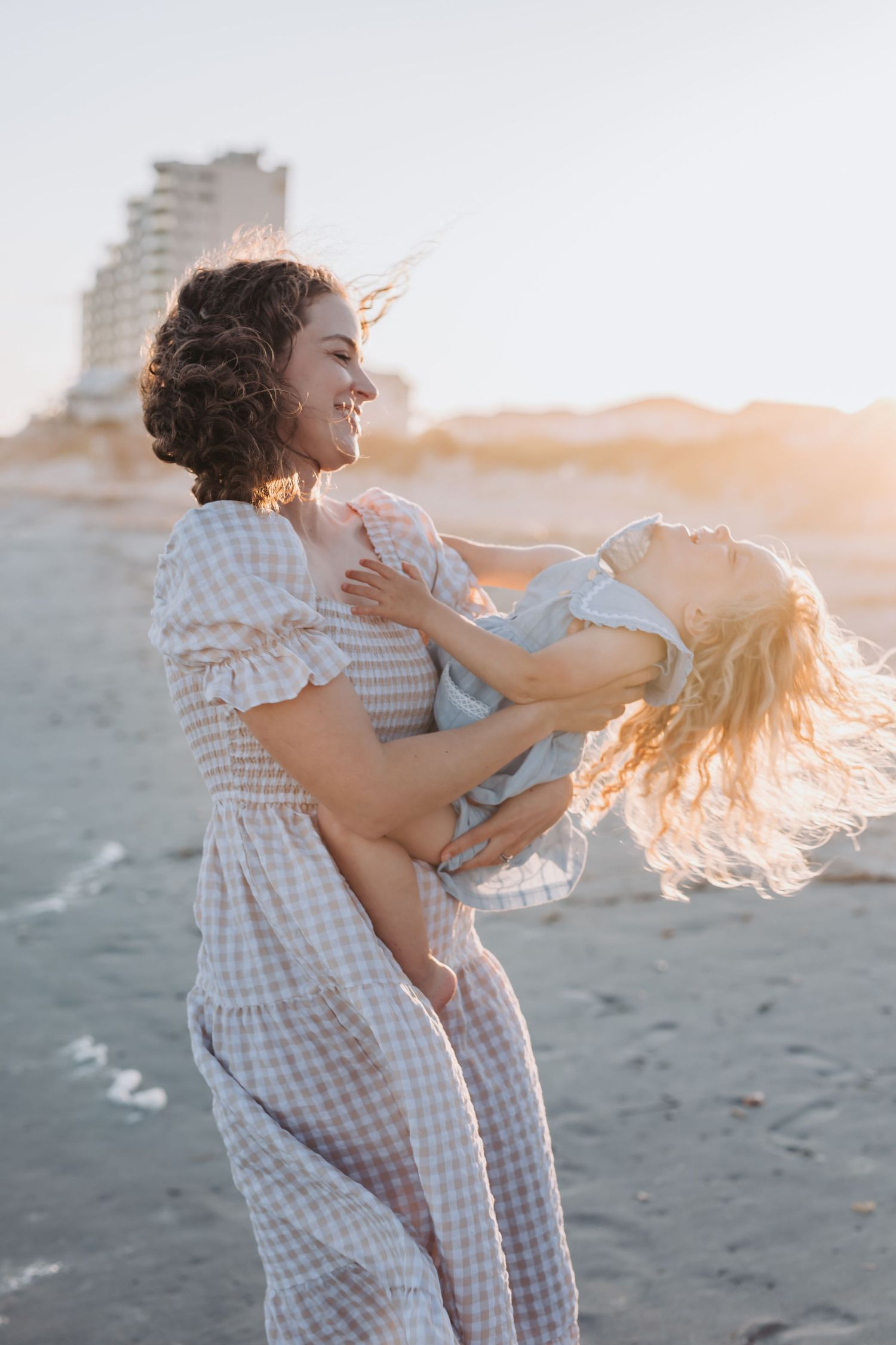 A mother and daughter in matching gingham dresses embrace and spin together on the beach at sunset.