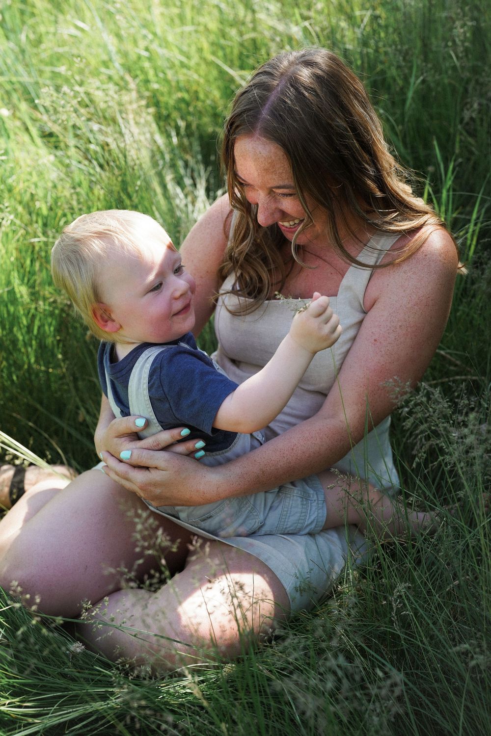 A joyful moment captured between a parent and child sitting together in tall green grass on a sunny day.