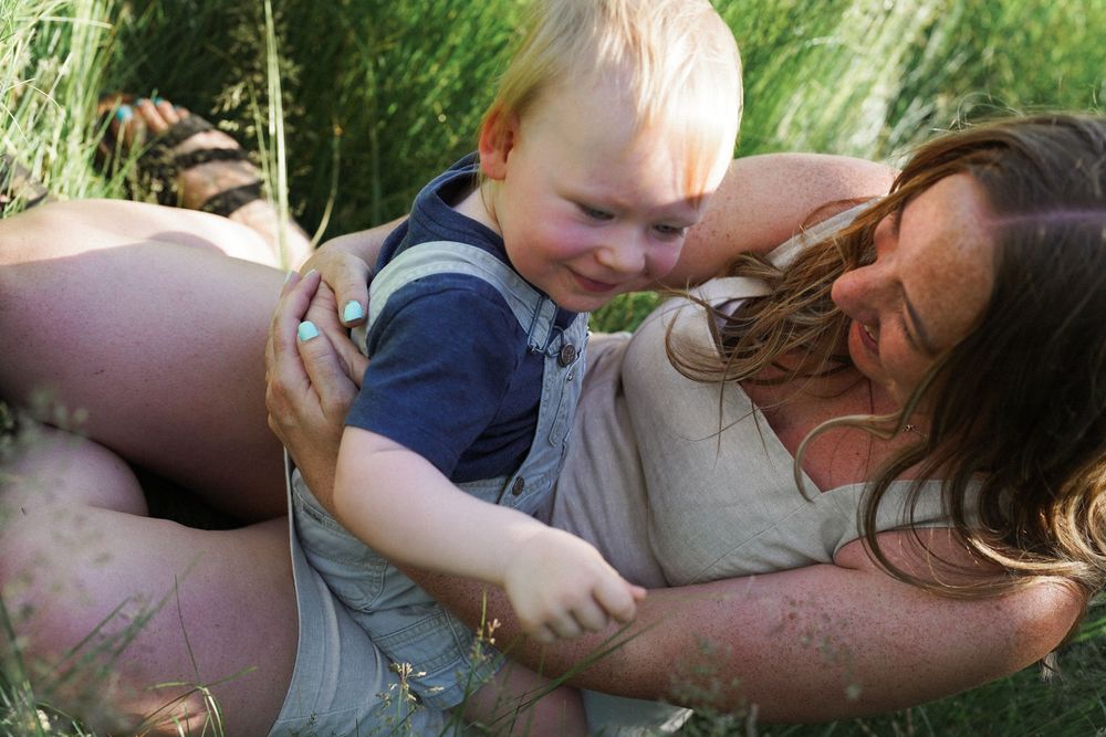 Young child in overalls plays and smiles joyfully while lying in green grass together with a caregiver.
