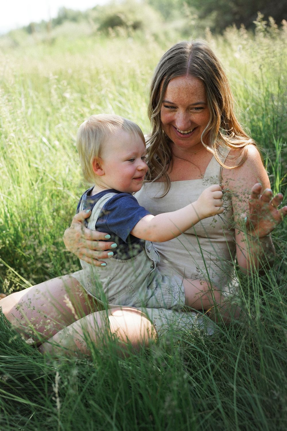 A parent and child share joyful moments together while sitting in tall summer grass at golden hour.
