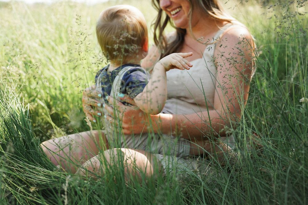 A mother wearing a lace dress sits with young child in tall grass during golden hour outdoor photography session.