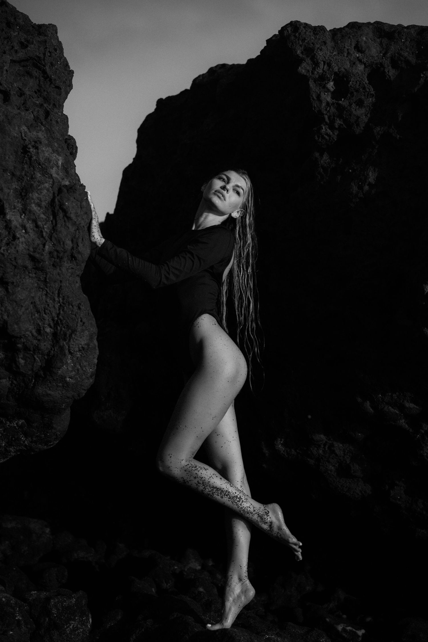 Artistic black and white photo of dancer posing elegantly against rocky cliff backdrop in dramatic lighting.