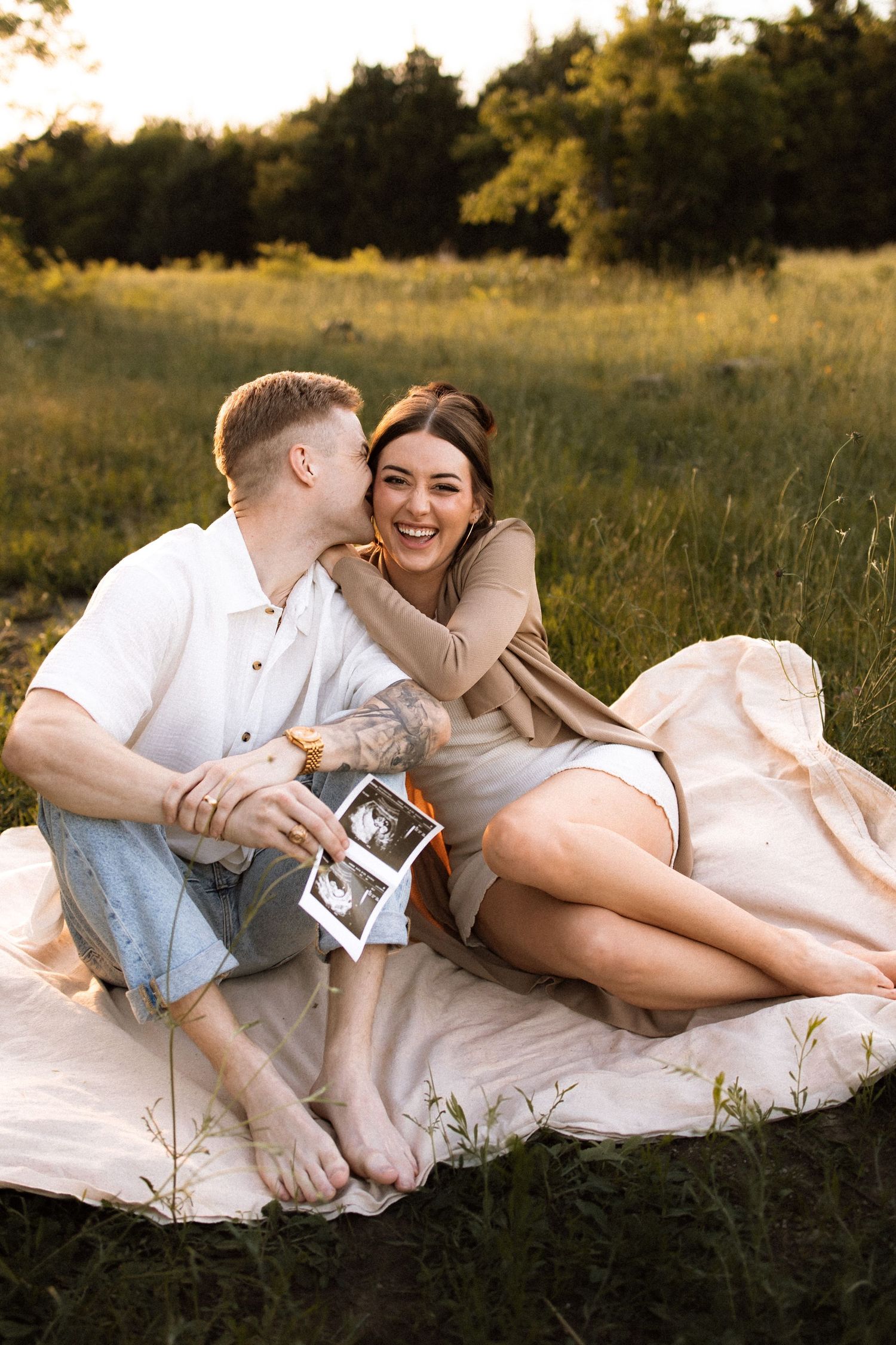 Couple standing on blanket in grassy area holding ultrasound photo while sharing romantic moment at sunset.