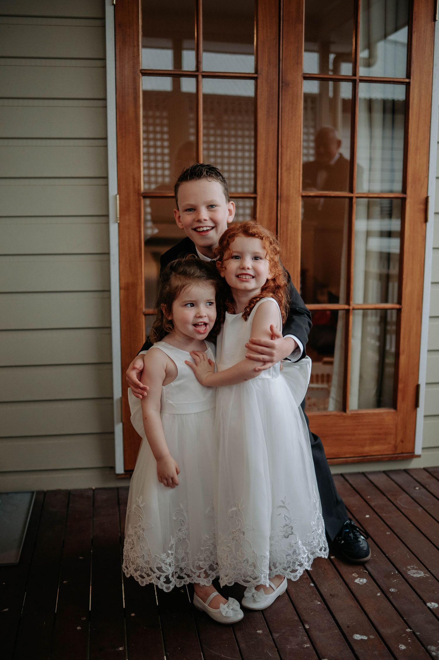 Three children in formal wear smile and embrace on a wooden porch in front of glass french doors.