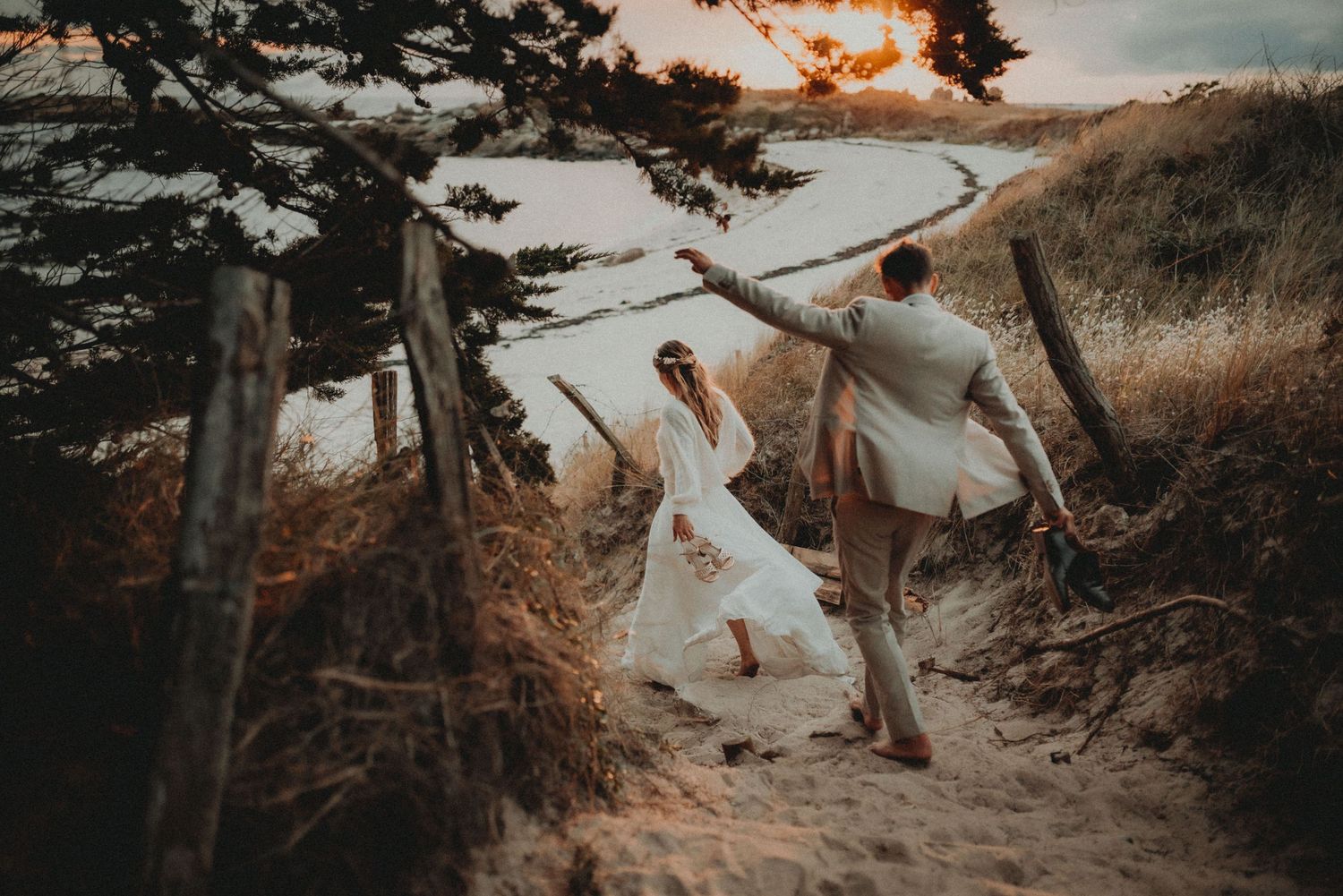 Playful wedding couple walking along scenic coastal path at sunset.