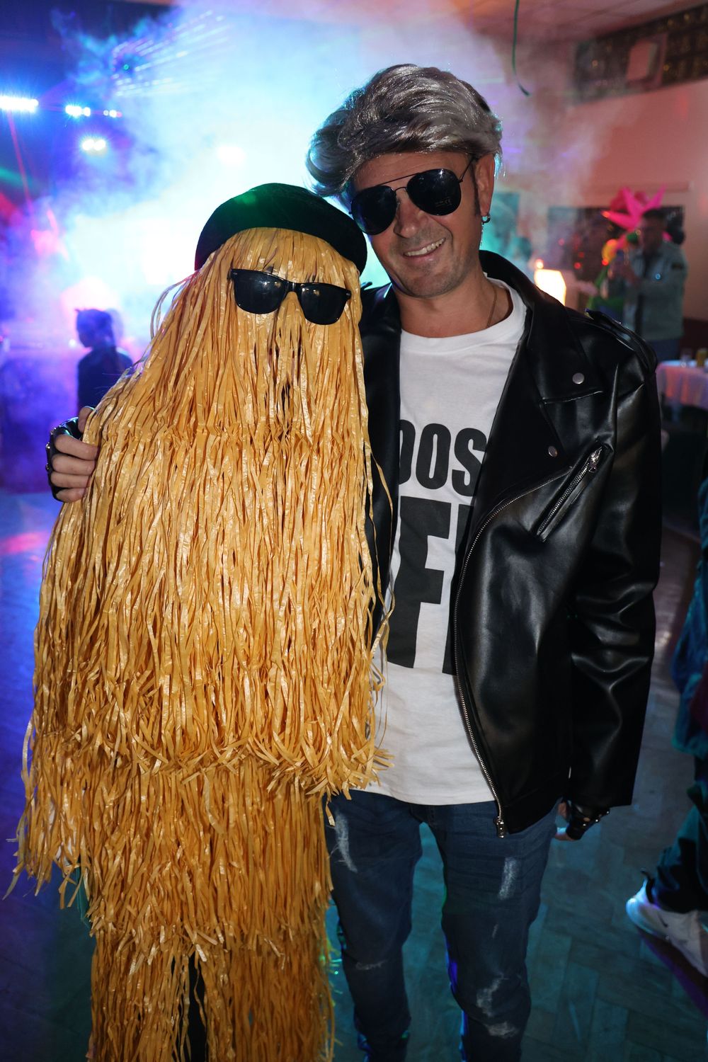 A person wearing a blonde straw-like wig and sunglasses poses at a party with colorful lighting in the background.