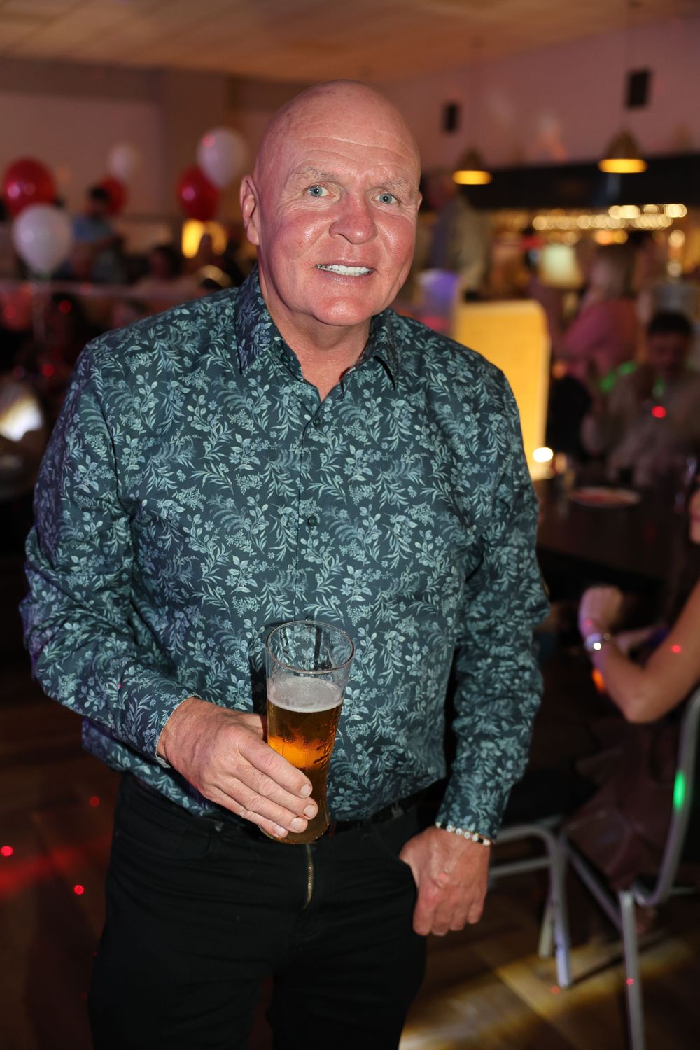 Person in a green patterned dress shirt holding a beer glass at a social gathering with ambient lighting in the background.