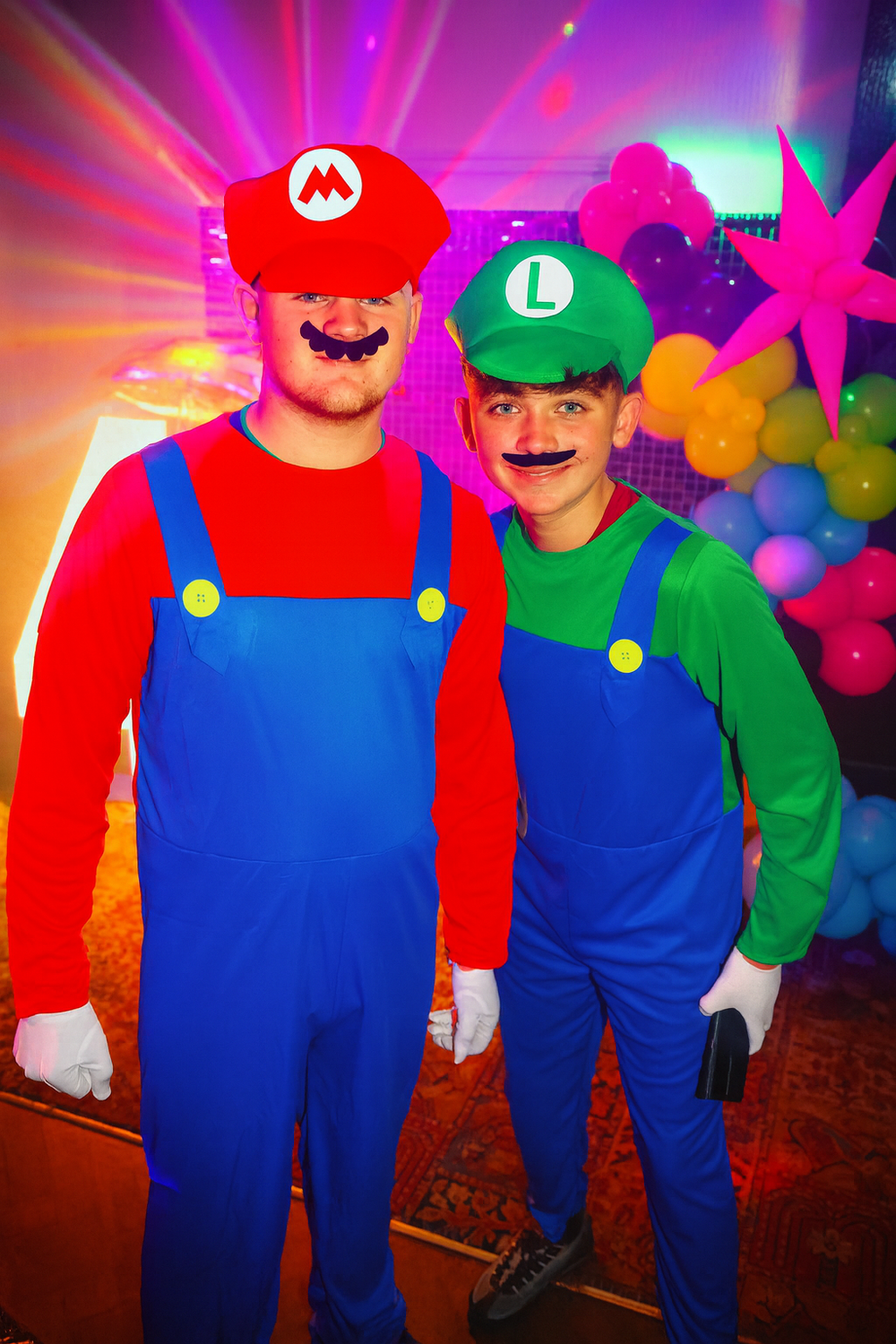 Two people dressed as Mario and Luigi from Nintendo's Super Mario Bros posing against colorful party decorations.