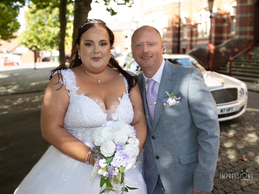 A couple in wedding attire poses for photos outdoors near brick buildings and trees on their special day.