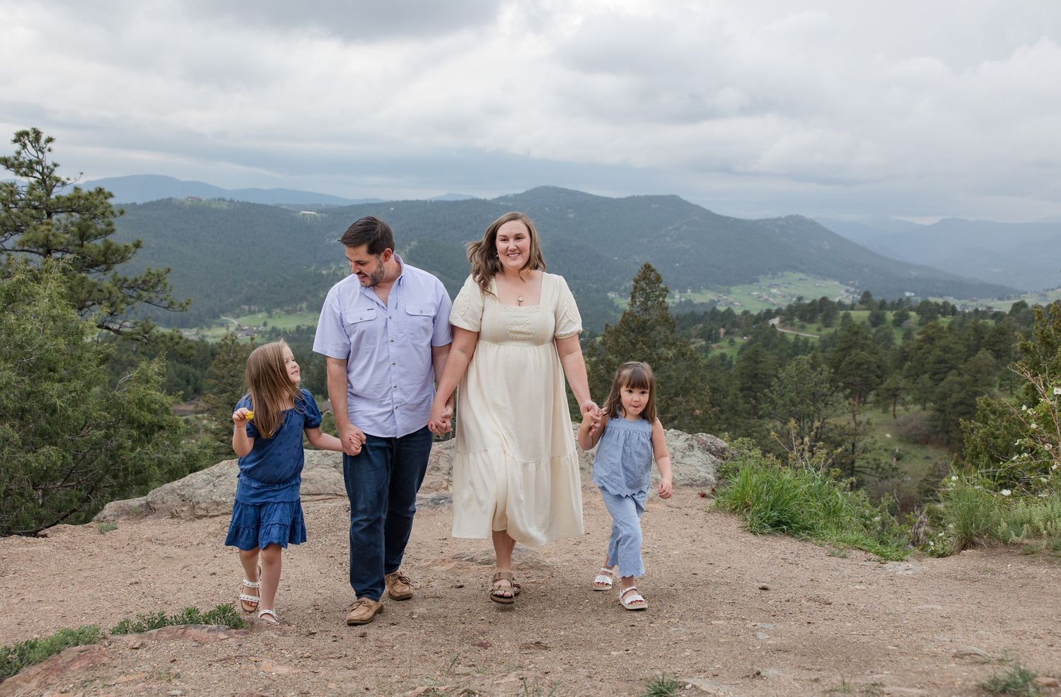 A family walks together along a mountain trail with scenic mountain ranges and forest in the background.