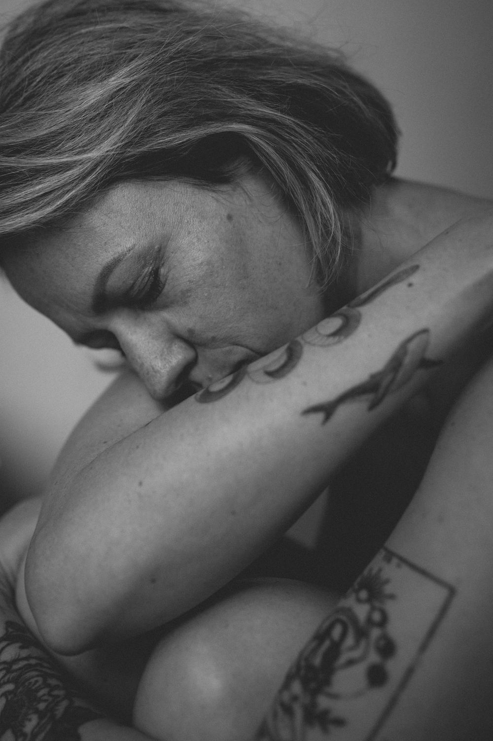 Black and white artistic photograph of sleeping figure with visible tattoos in soft, intimate lighting.