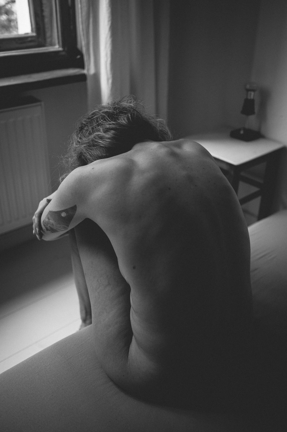 A black and white artistic nude photograph showing a person's bare back sitting by a window in moody lighting.