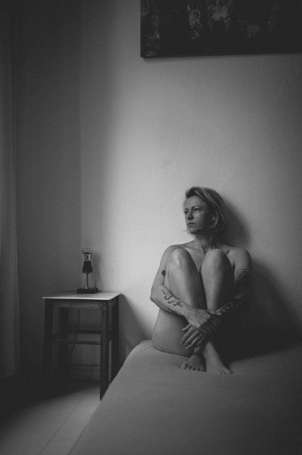 Artistic black and white boudoir photograph showing intimate bedroom scene with a glass bottle on bedside table.