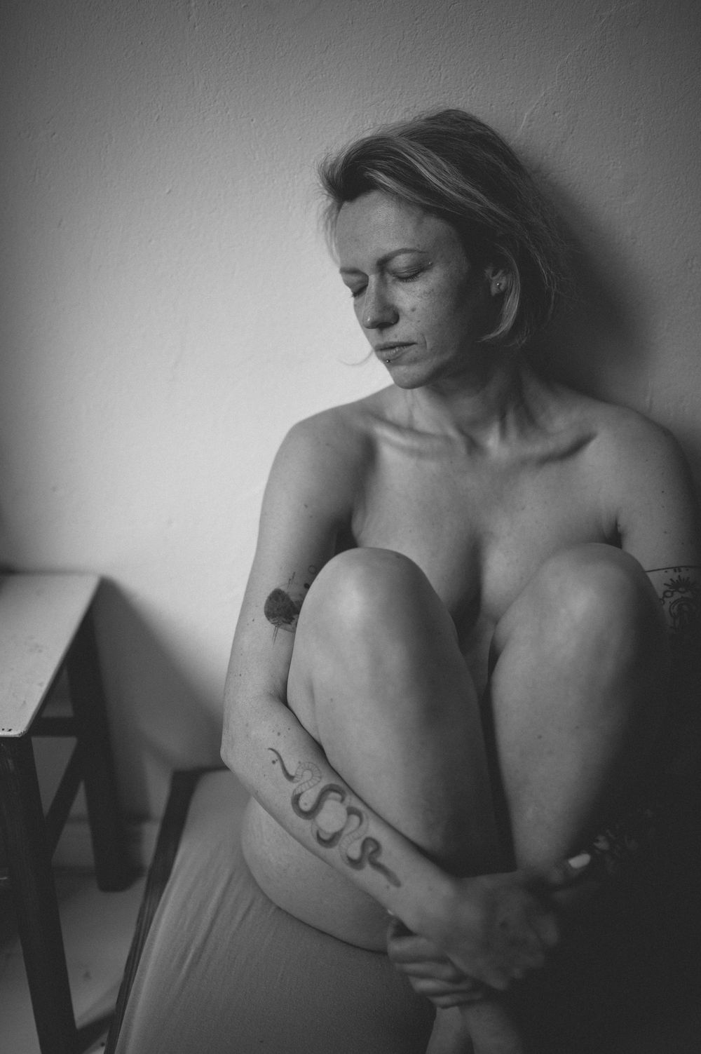 A black and white artistic portrait of someone seated in shadow with eyes closed, showing a snake tattoo on their leg.