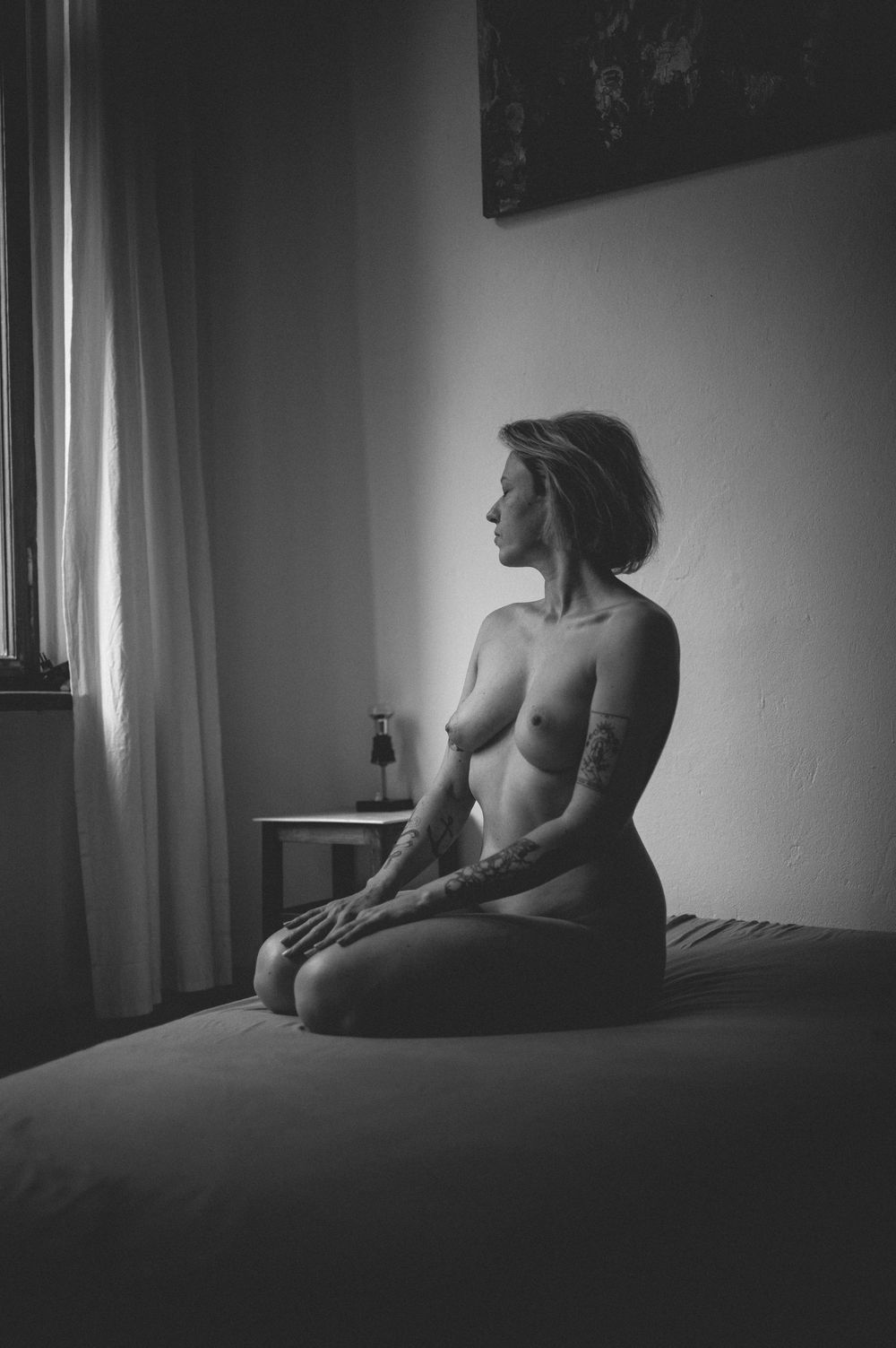 Black and white artistic photograph of a person sitting nude in profile on a bed near a window.