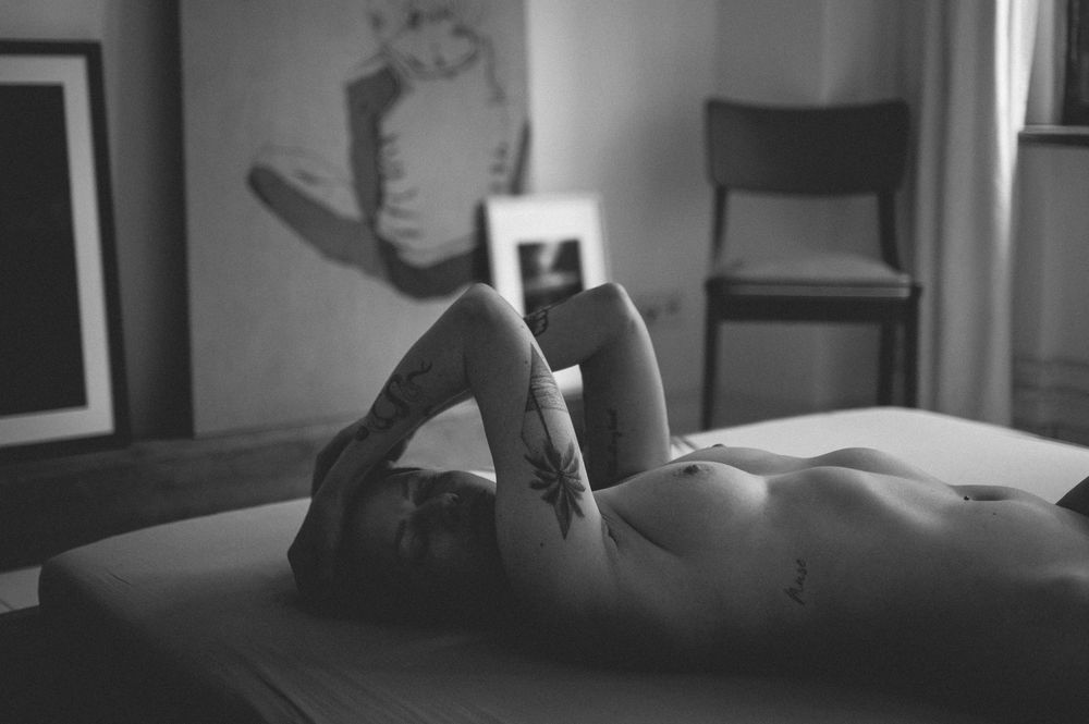 Black and white artistic nude figure study on bed with soft shadows and ambient lighting.