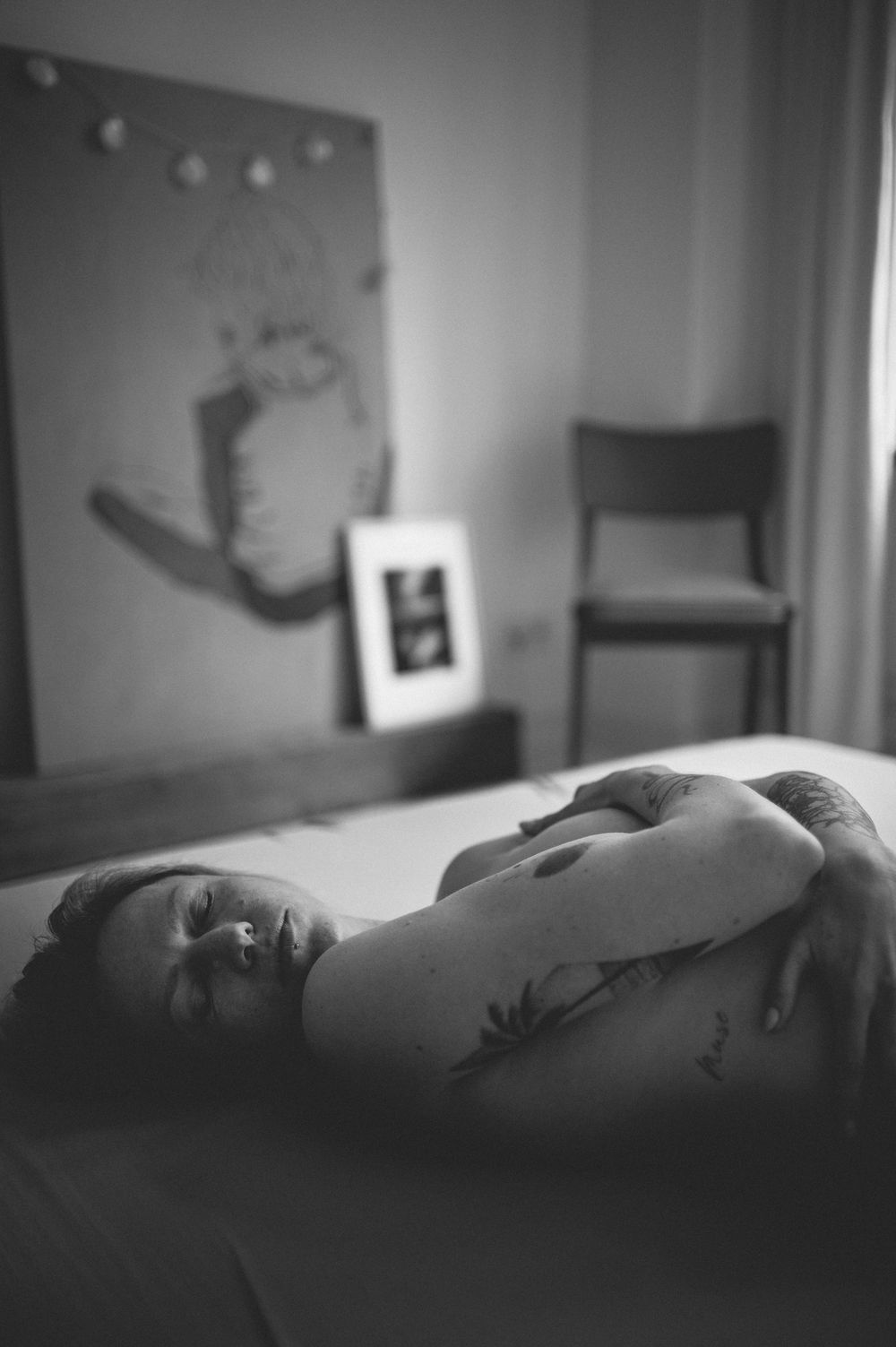 Artistic black and white boudoir photograph showing a reclining figure in dim bedroom lighting with wall art visible.