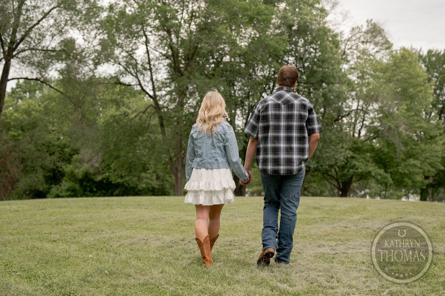 A couple holds hands while walking away from the camera across a grassy lawn on a sunny day.