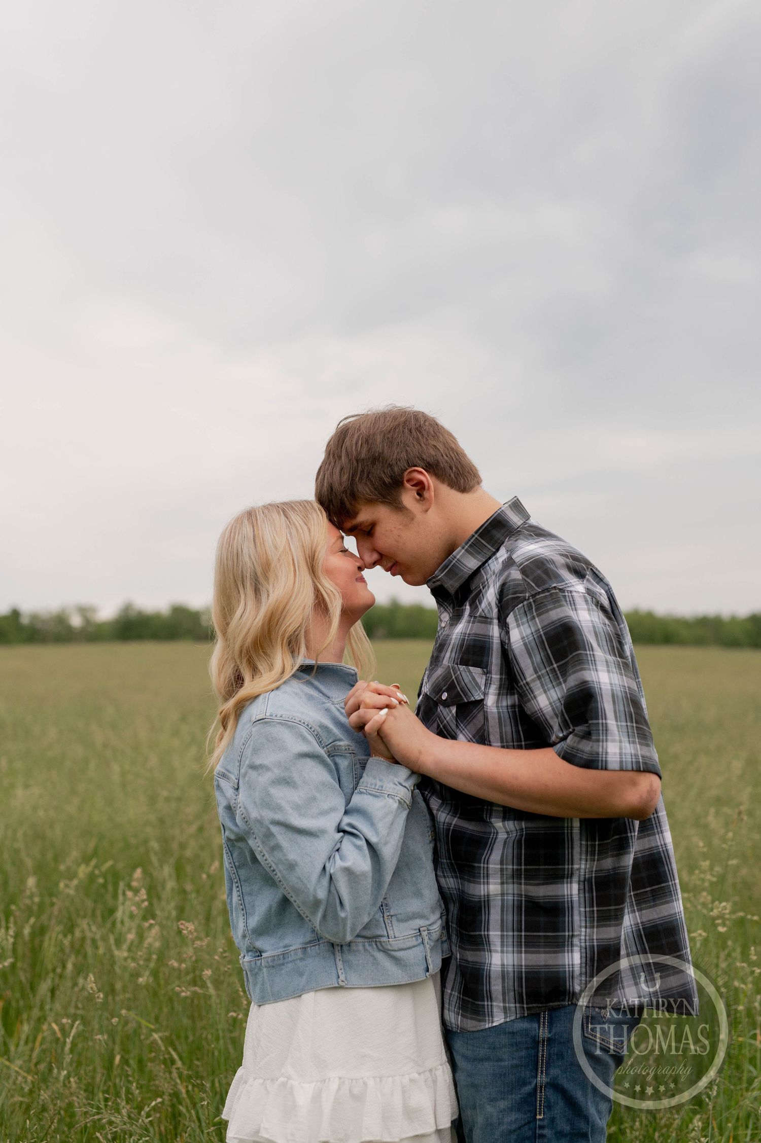 A romantic couple embraces in a serene grassy field on an overcast day, dressed casually in denim and plaid.
