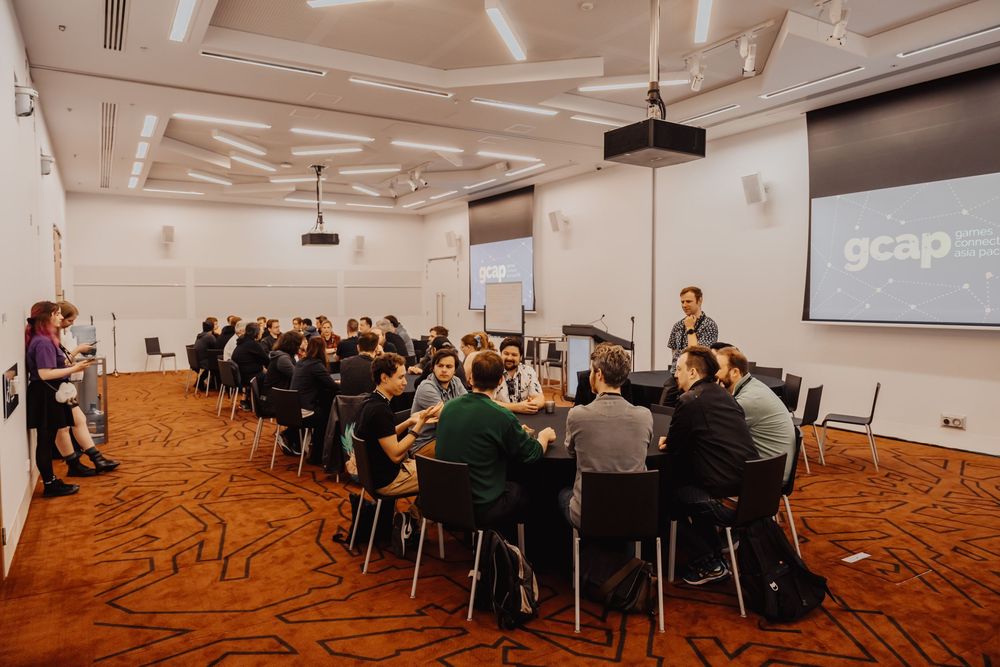Business professionals seated around tables in a modern conference room with geometric carpeting and projector screen.