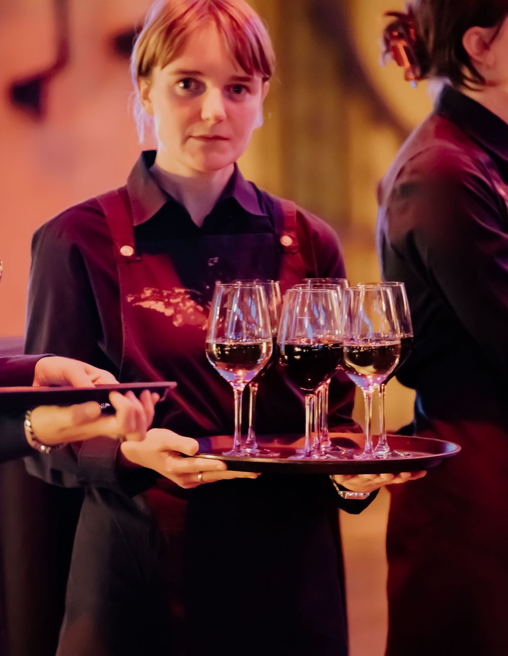 Server in burgundy apron carries a tray with multiple glasses of red wine in elegant restaurant setting.