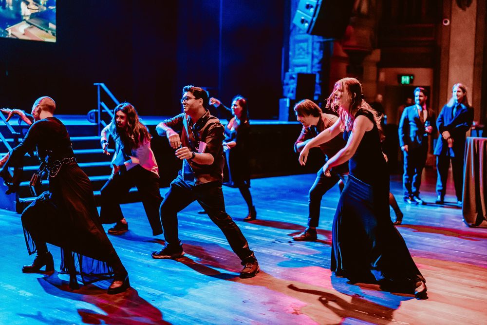 Dance class participants perform synchronized squatting movements on a wooden floor illuminated by blue stage lighting.