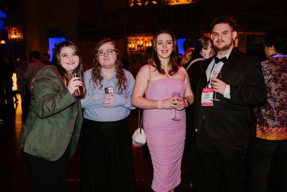 Four friends holding drinks pose together at an elegant evening event with mood lighting and decorative ambiance.