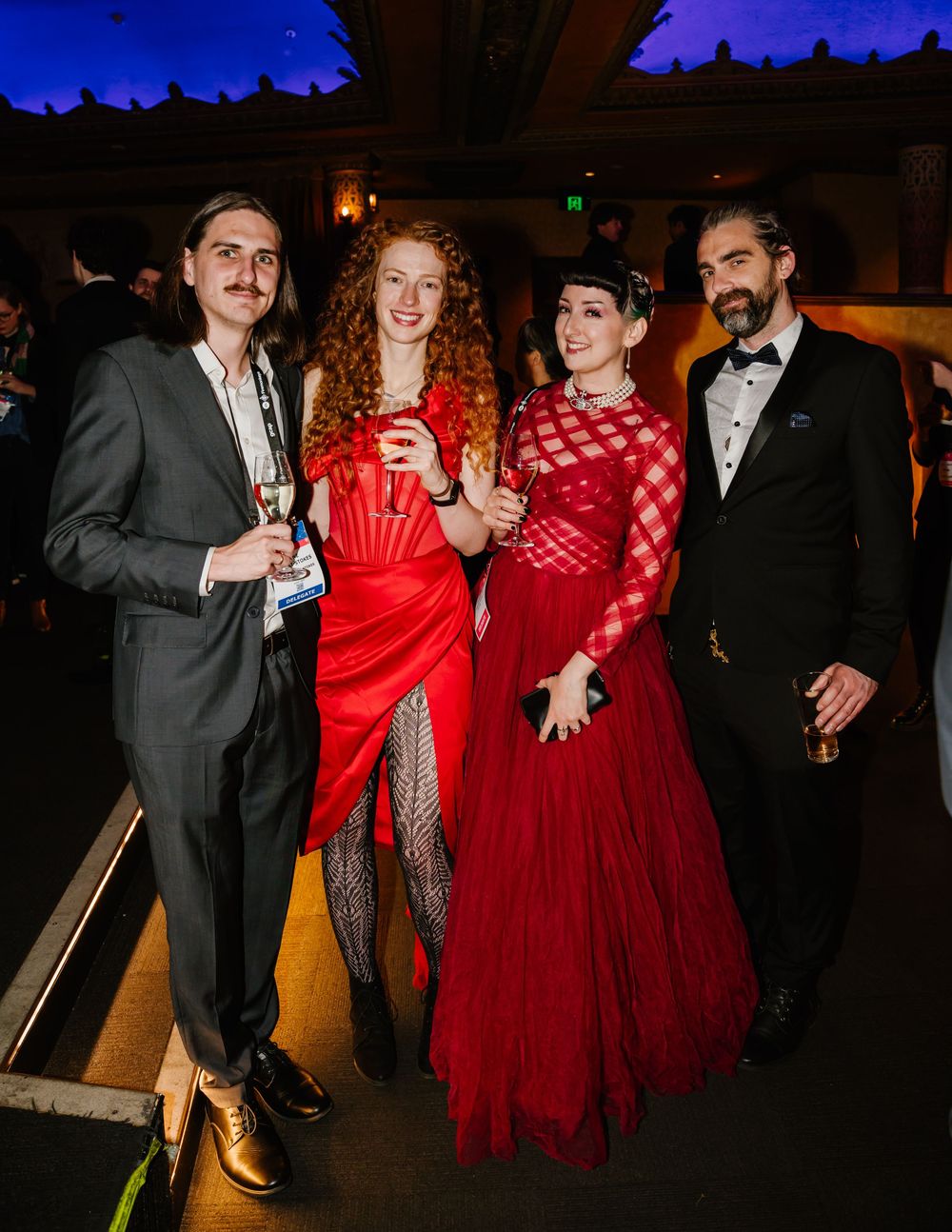 Elegant group in formal attire posing together at an evening event, with two in red gowns and two in black tuxedos.