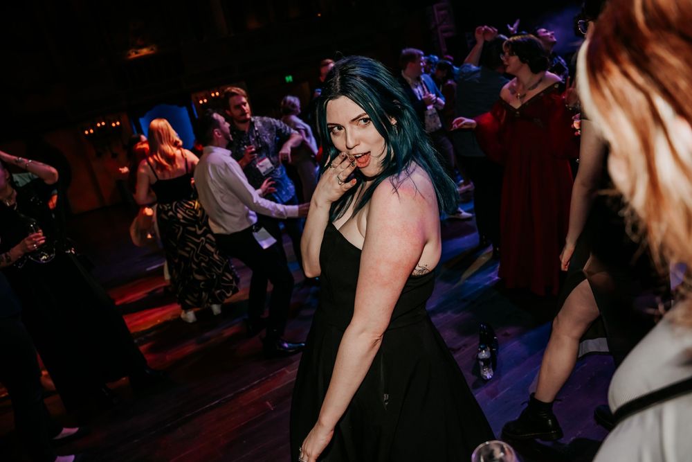 A woman in a black dress dances at a crowded nightclub with moody dark lighting and people in the background.
