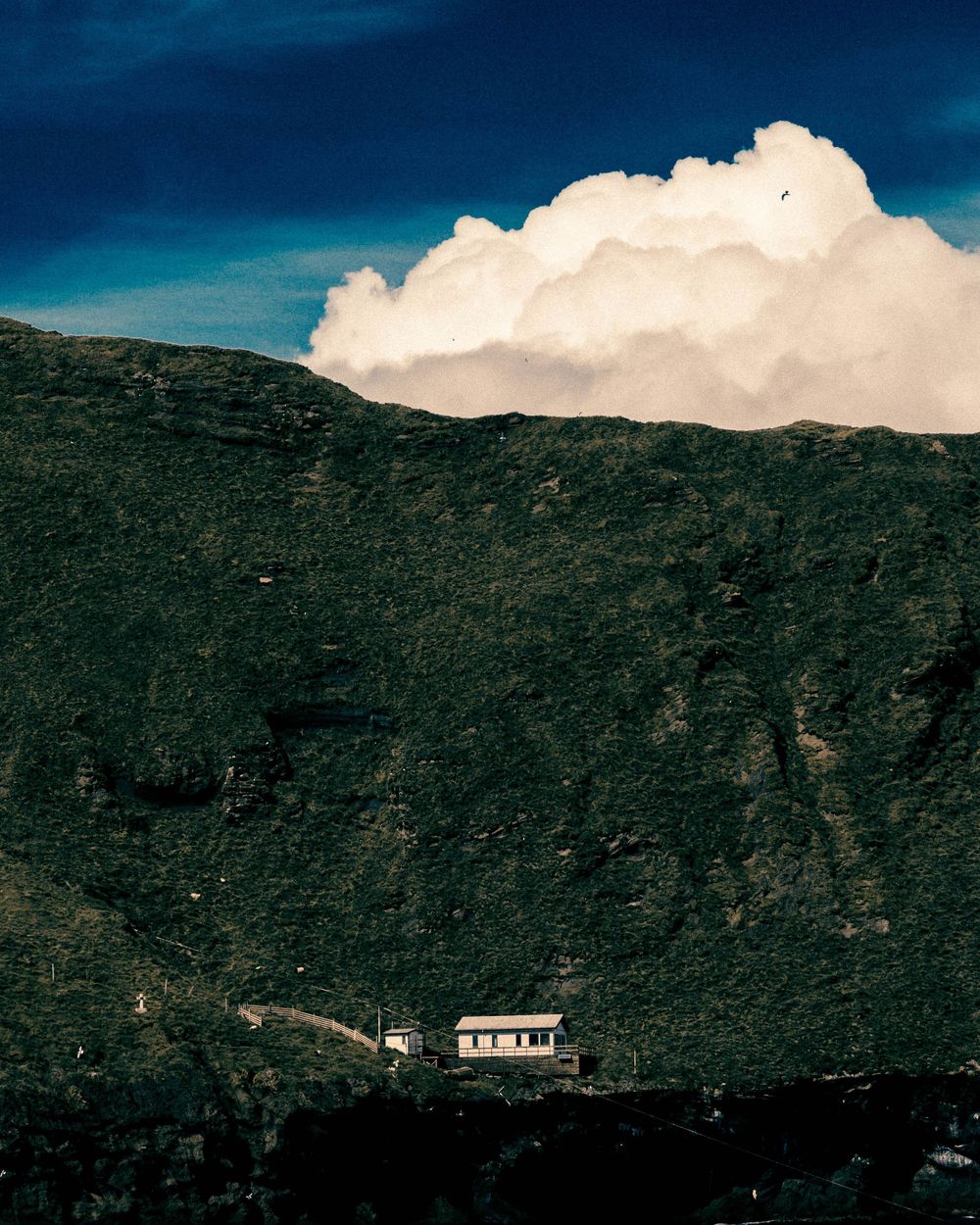 A small white house nestled on a lush green mountainside beneath dramatic white clouds and deep blue sky.