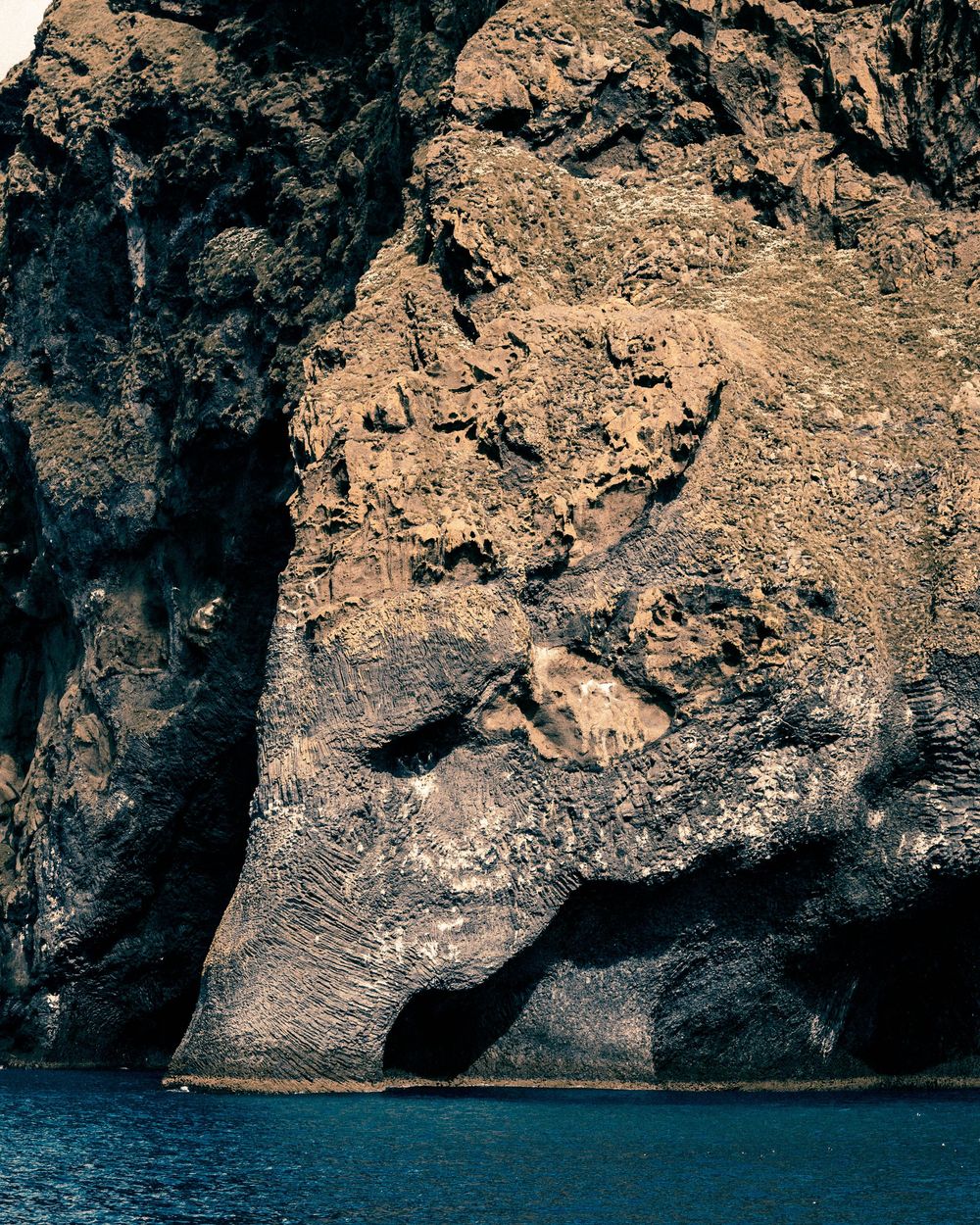 Dramatic rocky coastal cliffs meet deep blue waters with natural cave formations along a rugged shoreline.
