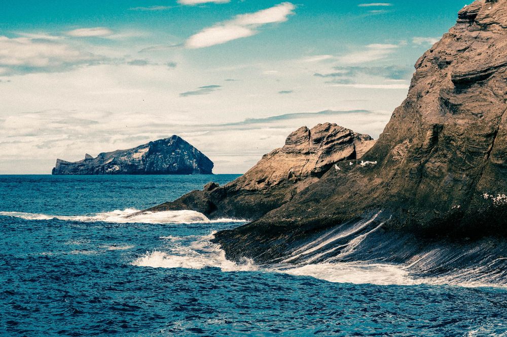 Rocky coastal cliffs rise dramatically above deep blue ocean waters with waves crashing against the rugged shoreline.