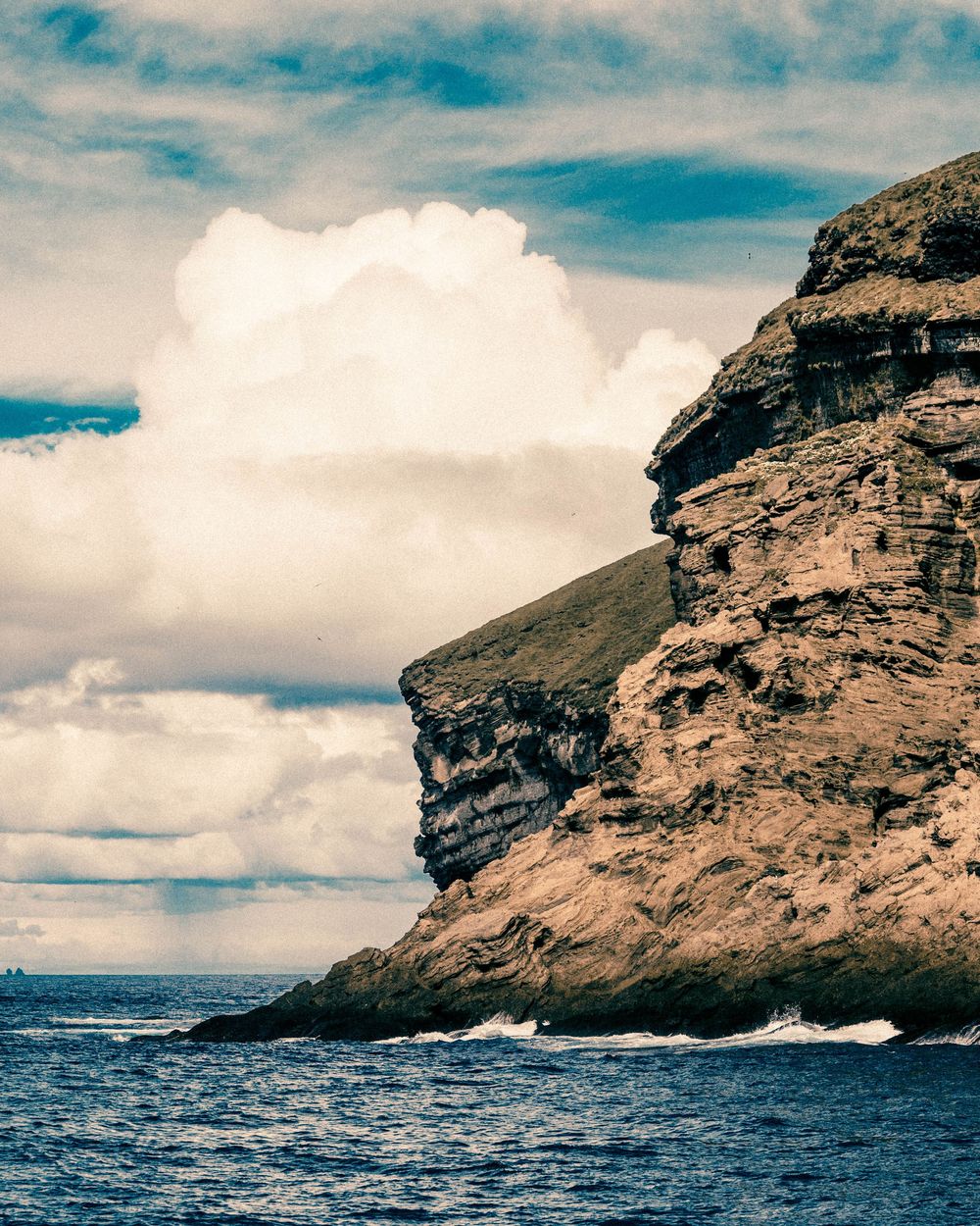 Dramatic rocky coastal cliff rises above turbulent ocean waves under a cloudy blue sky with white puffy clouds.