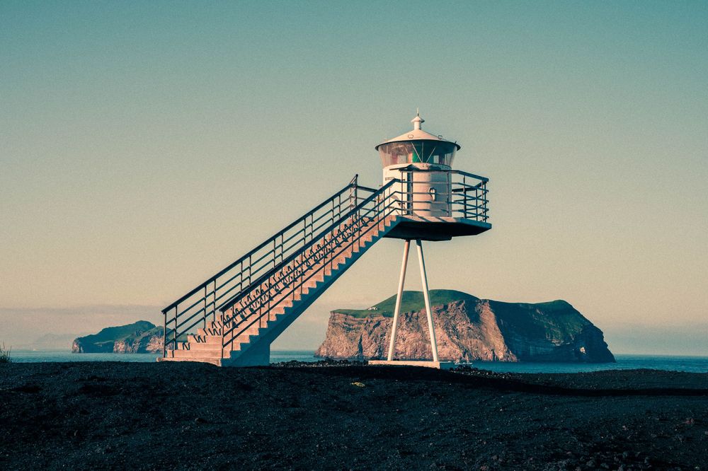 A vintage lifeguard tower with stairs overlooks a dramatic coastal landscape at sunset with rocky cliffs in the distance.