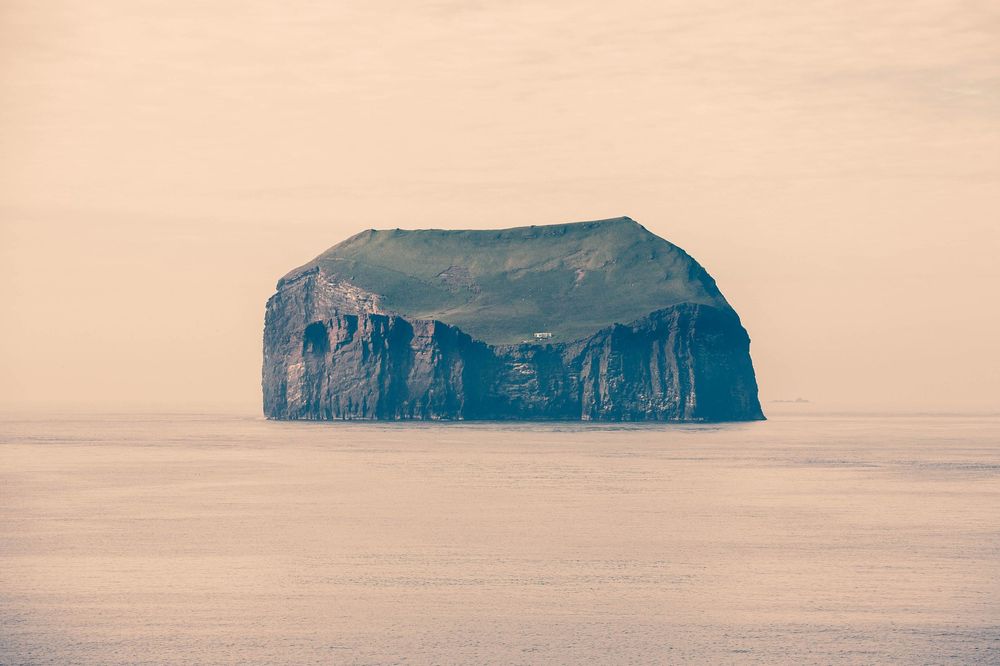 A solitary rocky island with steep cliffs rises from misty waters during golden hour.